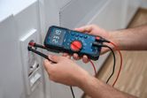 Oseba uporablja digitalni multimeter za preverjanje napetosti v stenski vtičnici.