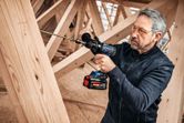 Oseba, ki nosi varnostno opremo, z akumulatorskim vrtalnikom privije vijak v lesene tramove.