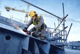 Oseba, ki nosi varnostno opremo, brusi velik kovinski tram s kotnim brusilnikom.