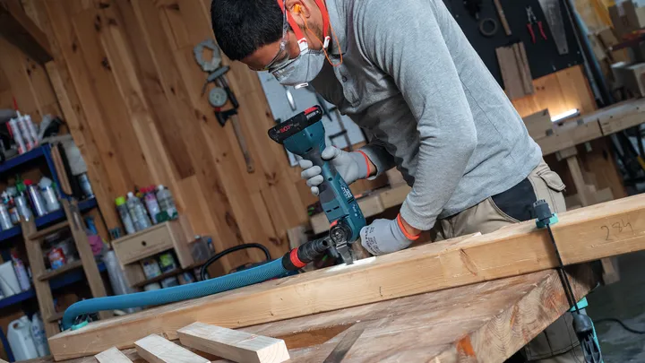 Oseba v varnostni opremi vrta v lesen tram na delovni mizi.