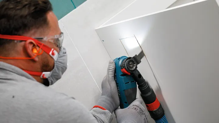 Oseba, ki nosi varnostno opremo, z električnim orodjem izreže pravokotno luknjo v plošči.