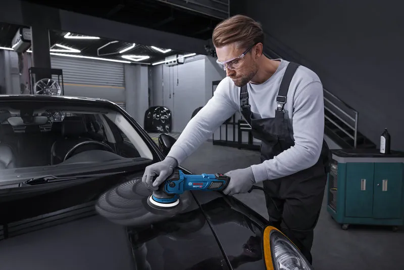 Oseba v varnostni opremi polira črn pokrov motorja avtomobila s polirno krtačo.