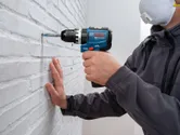 Oseba v varnostni opremi vrta v opečno steno z akumulatorskim udarnim vrtalnikom.