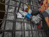Oseba, ki nosi varnostno opremo, uporablja akumulatorsko orodje za rezanje armature na betonski površini.
