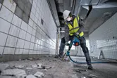 Oseba, ki nosi varnostno opremo, uporablja vrtalno kladivo za razbijanje ploščic na tleh.