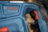 Oseba, ki nosi varnostno opremo, drži moder ročaj električnega orodja z rdečim sprožilcem.