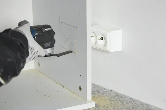Osoba v bezpečnostnom vybavení vyreže štvorcový otvor v drevenom paneli pomocou elektrického náradia.