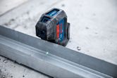 Laserový nivelačný nástroj premietajúci zelené laserové čiary pozdĺž kovovej dráhy na betónovej podlahe.