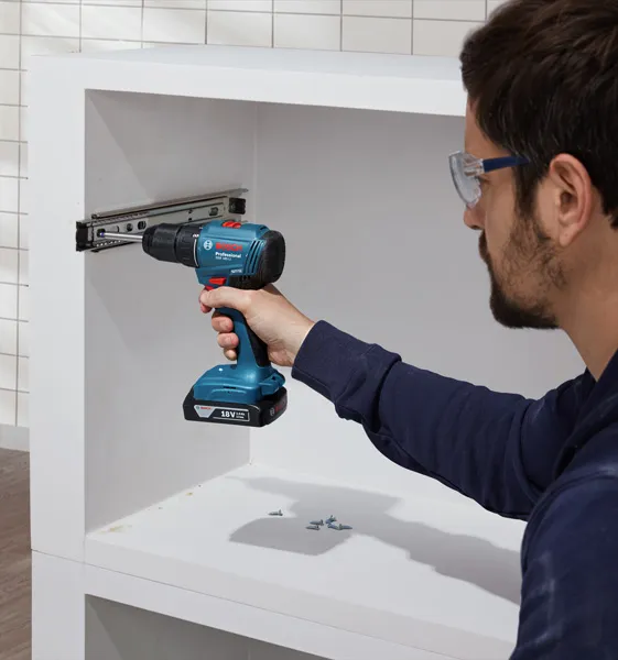 Osoba v bezpečnostnom vybavení používa akumulátorovú vŕtačku na inštaláciu výsuvov zásuviek do skrinky.
