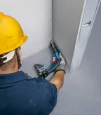 Osoba v bezpečnostnom vybavení zaskrutkuje skrutky do sadrokartónu pomocou elektrického náradia.