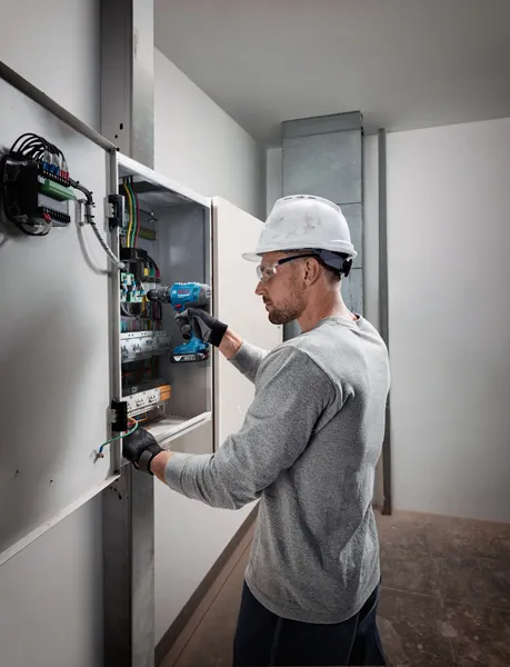 Osoba s ochranným vybavením používa akumulátorovú vŕtačku na elektrickom paneli.