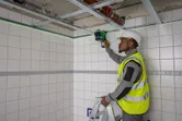 Osoba v bezpečnostnom vybavení nastavuje laserový nivelačný prístroj na obloženú stenu z rebríka.