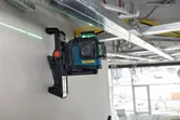 Laserový nivelačný prístroj namontovaný na stene, ktorý premieta zelený laserový lúč cez stavenisko.