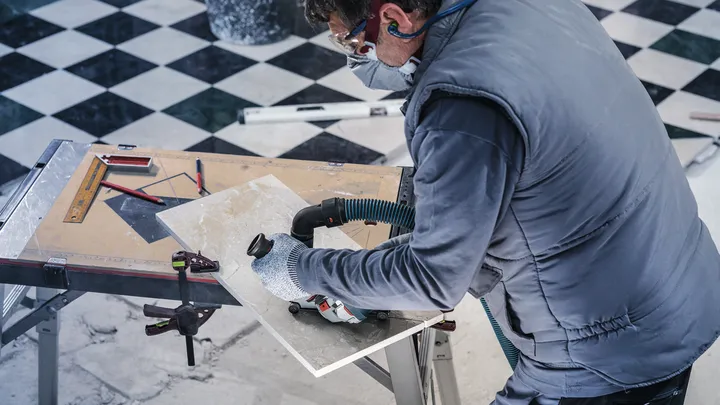 Osoba v bezpečnostnom vybavení reže dlaždice elektrickým náradím na pracovnom stole.