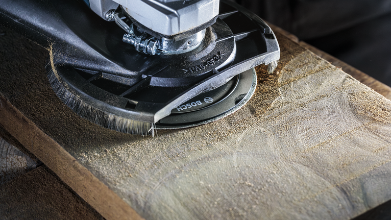 Uhlová brúska s nadstavcom z drôtenej kefy vyhladzuje povrch drevenej dosky.