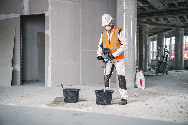Osoba s ochranným vybavením používa miešací mechanizmus na miešanie materiálu vo vedre.