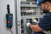 Osoba v bezpečnostnom vybavení testuje elektrický panel pomocou digitálneho multimetra.