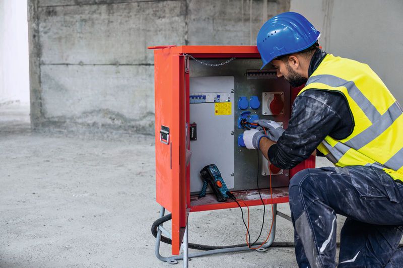 Osoba s bezpečnostným vybavením používa digitálny multimeter na testovanie elektrického panela.