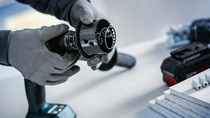 Osoba v bezpečnostnom vybavení pripevňuje dierovú pílu na vŕtačku v blízkosti naskladaných dosiek.