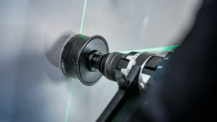 Osoba používa elektrickú vŕtačku s dierovou pílou na rezanie dlaždíc, pričom ju riadia zelené laserové čiary.