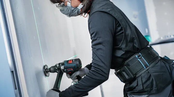 Osoba v bezpečnostnom vybavení vŕta do steny pomocou zeleného laserového lúča.