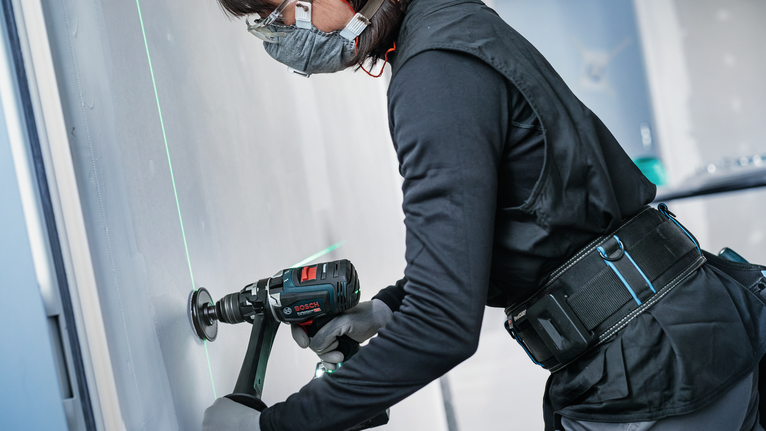 Osoba v bezpečnostnom vybavení vŕta do steny pomocou zeleného laserového lúča.