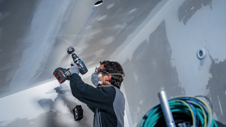 Osoba v bezpečnostnom vybavení vŕta do stropu na stavenisku.