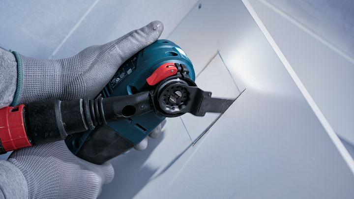 Osoba v bezpečnostnom vybavení vyreže obdĺžnikový otvor v sadrokartóne pomocou elektrického náradia.