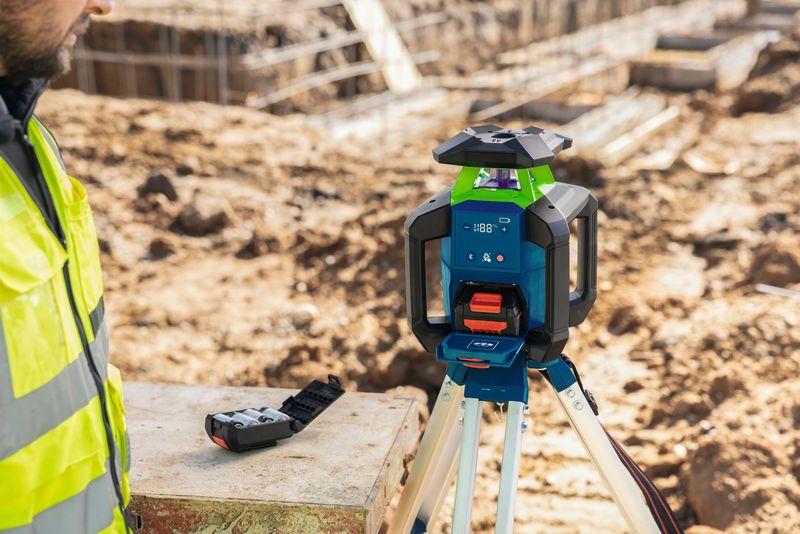 Osoba v bezpečnostnom vybavení pozoruje laserový nivelačný prístroj umiestnený na statíve.