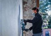 Osoba v bezpečnostnom vybavení obsluhuje drážkovaciu frézu na betónovej stene.