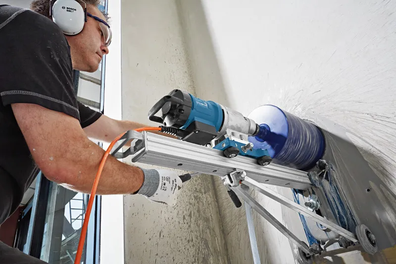 A person wearing safety equipment operates a drill stand to core a hole in a concrete wall.