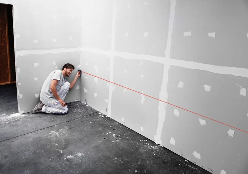 Person uses a laser measure against drywall, projecting a straight red guide line.