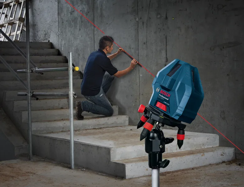 A person marks a concrete wall near stairs, guided by a laser leveling tool.