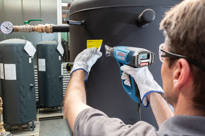 A person wearing safety equipment uses a heat gun to apply a label on a large tank.
