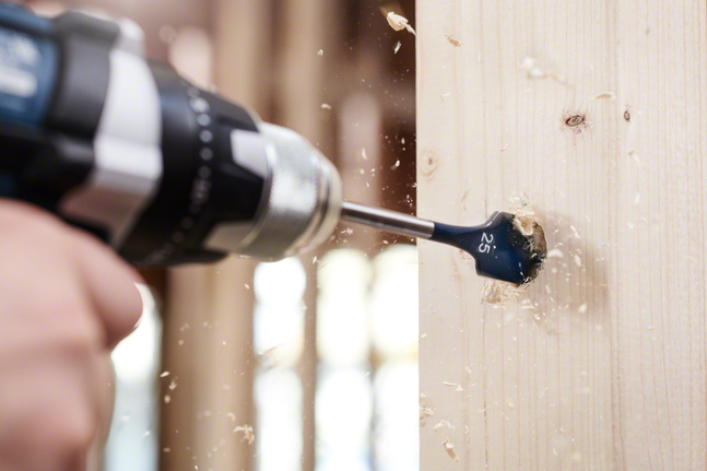 Person drilling a large hole in wood with sawdust flying.