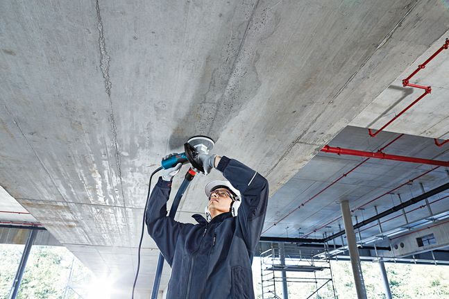 A person wearing safety equipment grinds a concrete ceiling with a power tool.