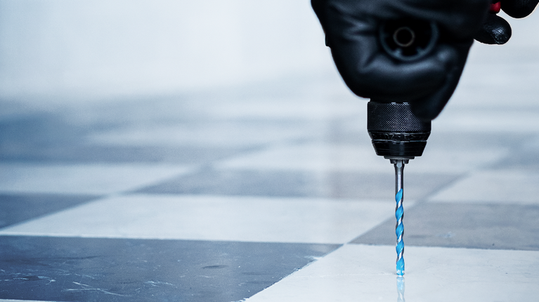 Person wearing safety equipment drills into a tiled floor using a power tool.