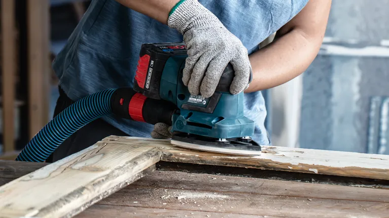 Worker wearing safety equipment sands wooden planks with a handheld power sander.