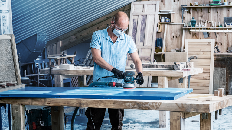 Person wearing safety equipment sands a blue door on a wooden workbench.