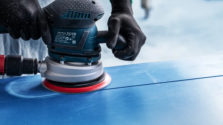 Person wearing safety equipment sands a blue surface with a power sander.