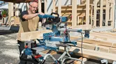 A person wearing safety equipment cuts wood beams with a sliding mitre saw at a construction site.