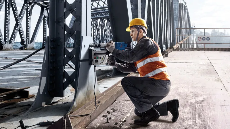 A person wearing safety equipment drills into a steel beam on a bridge.