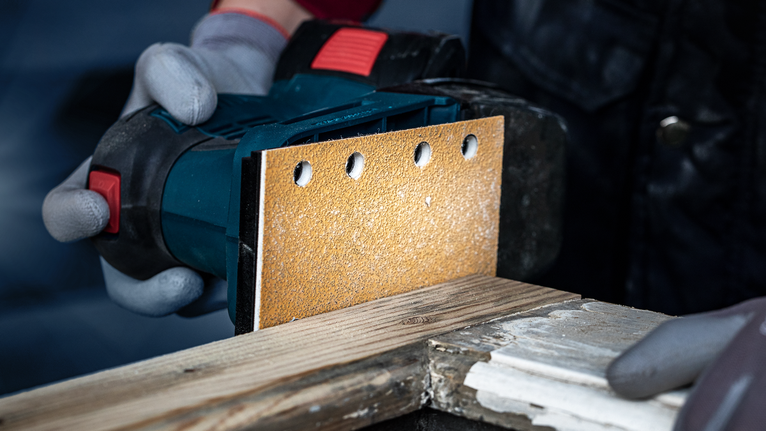 A worker wearing safety equipment sands a wooden plank with a handheld power sander.