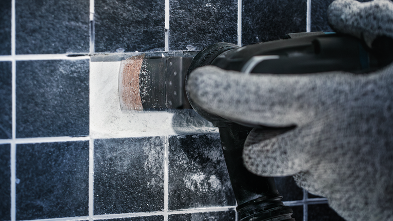 Gloved worker cutting a rectangular opening in tile using a power tool.