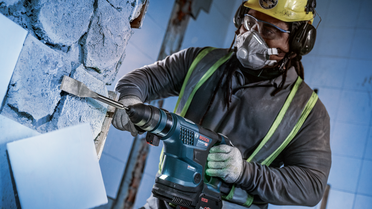 Person wearing safety equipment uses rotary hammer to chisel tiles off a wall.