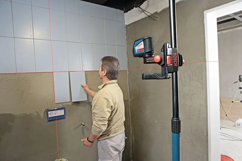 A person aligns wall tiles with a laser leveling tool mounted on a telescopic pole.