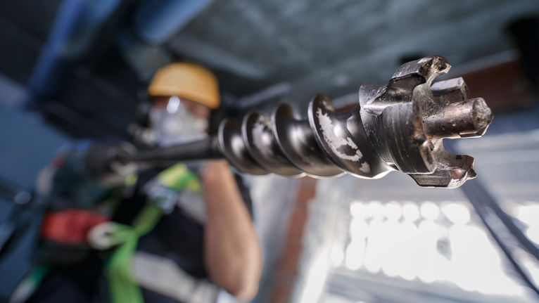 Person wearing safety equipment operates a large drill bit indoors.