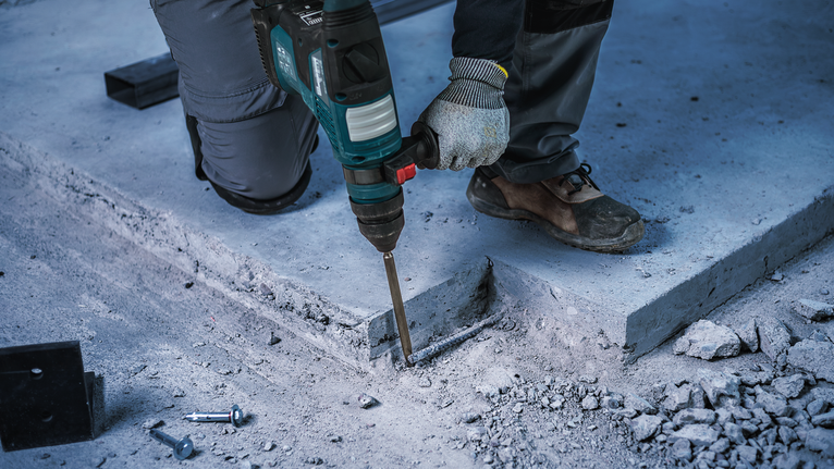 A worker wearing safety equipment uses a demolition hammer to break concrete.
