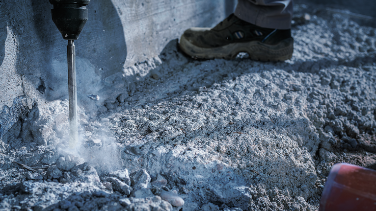 Worker wearing safety equipment drills into concrete at a construction site.