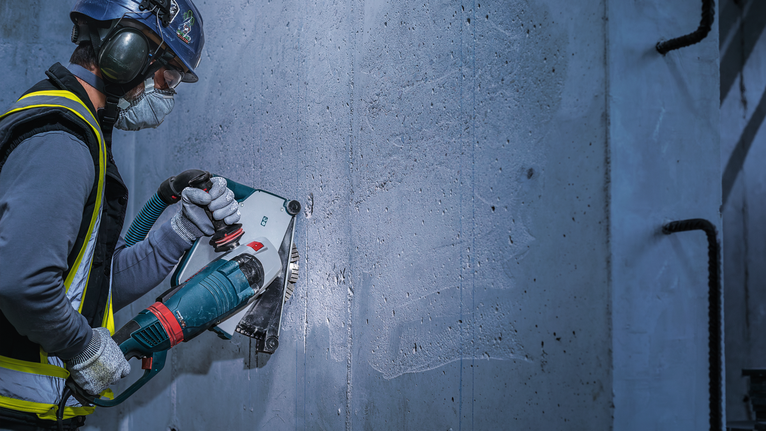 Person wearing safety equipment uses a power tool to cut into a concrete wall.